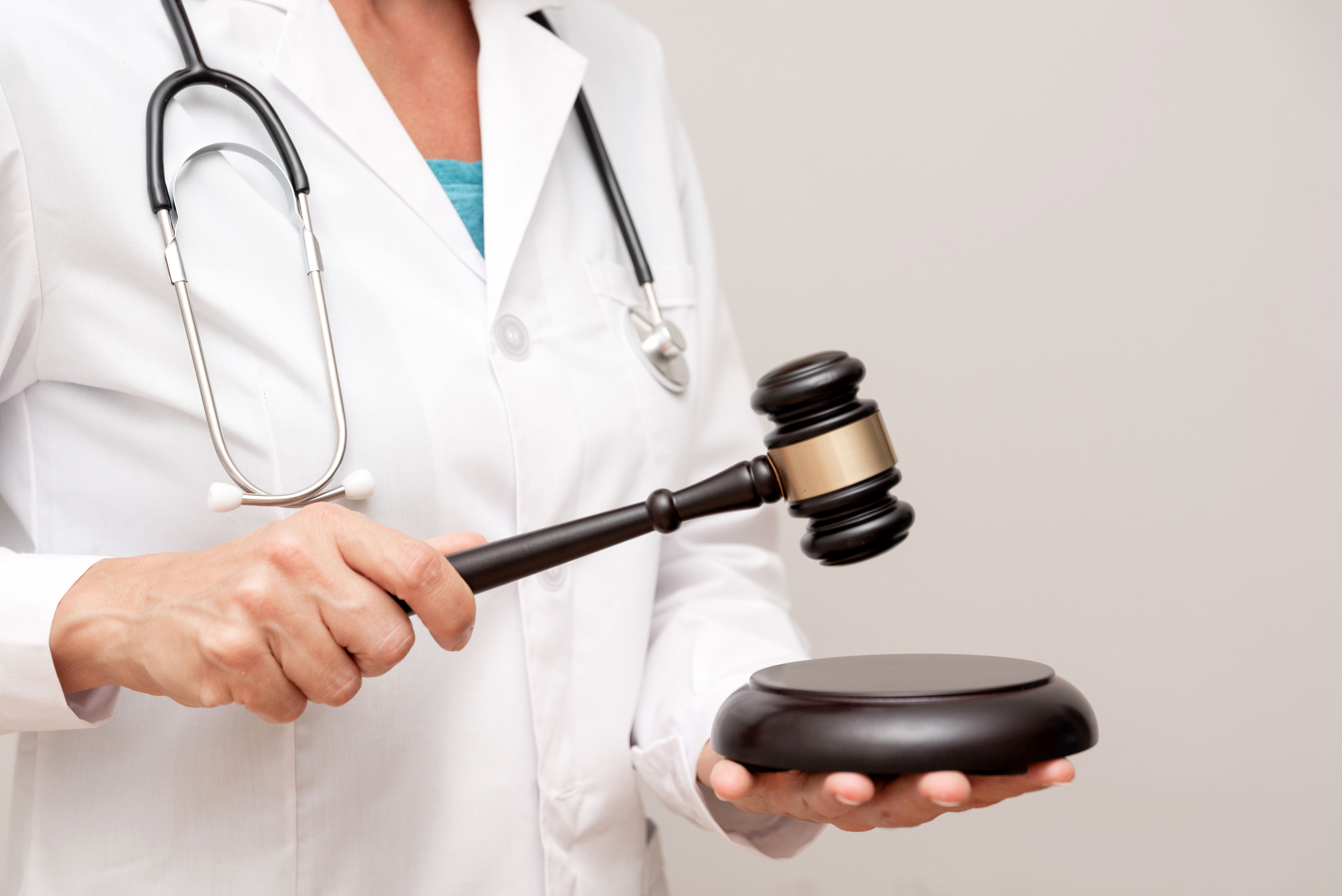 The torso of a doctor in a white jacket with a stethoscope over her shoulders. She is holding a gavel and sound block.