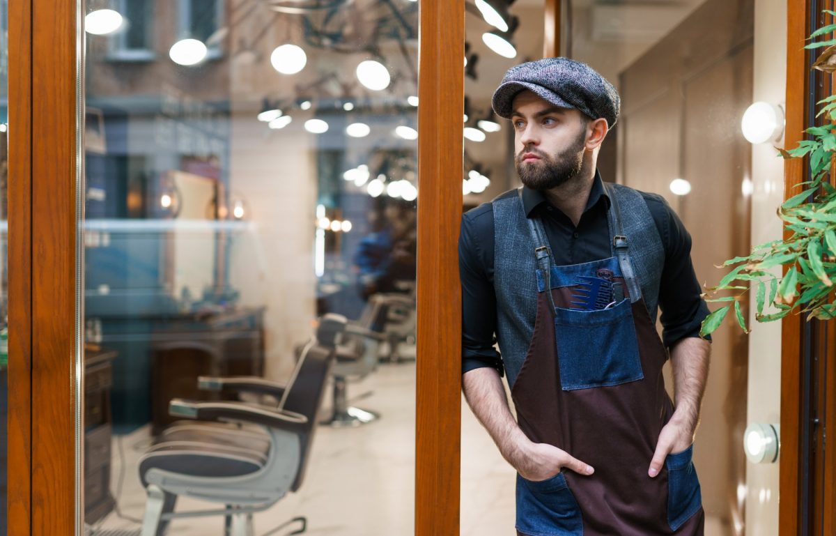 A man in a blue apron, blue hat, blue vest, and black shirt stands at the entrance of a business with barbershop chairs.