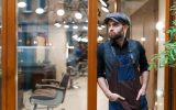 A man in a blue apron, blue hat, blue vest, and black shirt stands at the entrance of a business with barbershop chairs.