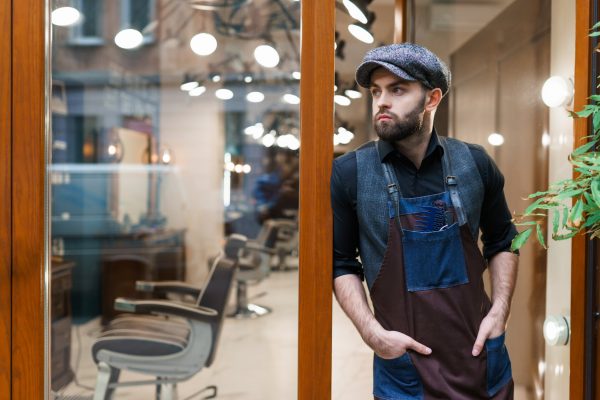 A man in a blue apron, blue hat, blue vest, and black shirt stands at the entrance of a business with barbershop chairs.