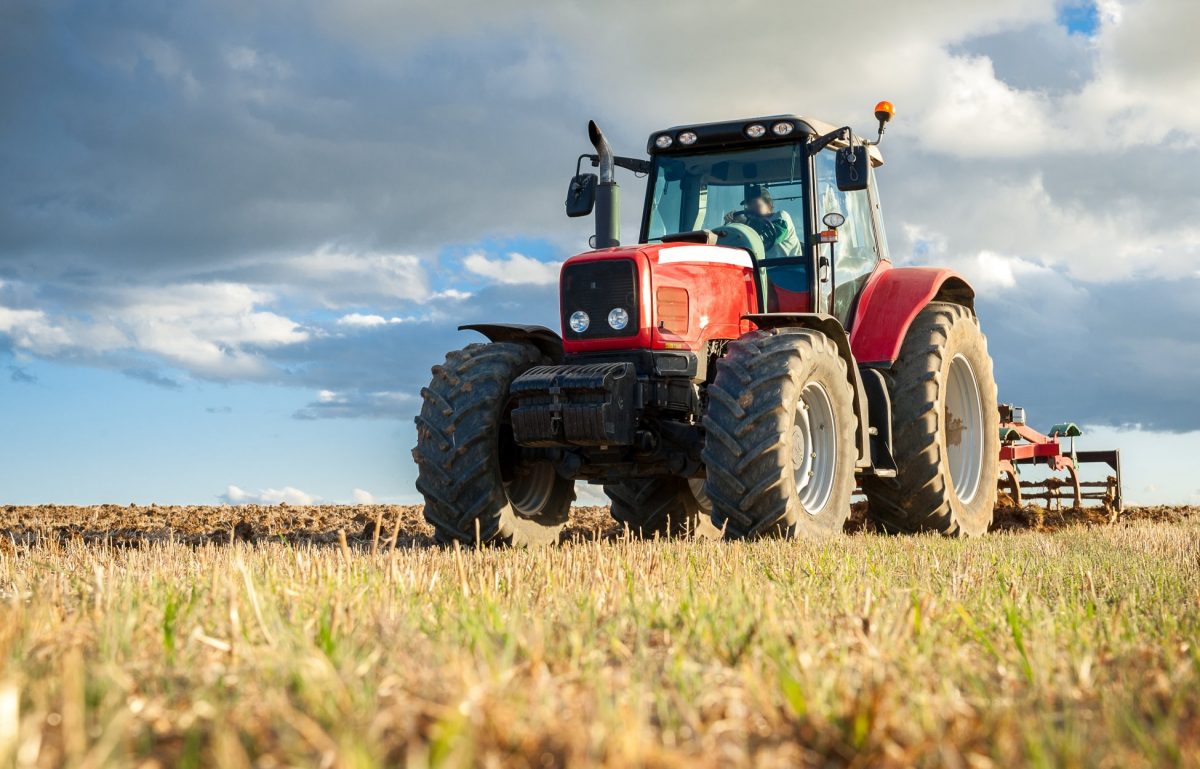 A person sitting in the cab of a red tractor in an open field. Though the sky is full of clouds, the sun shines.