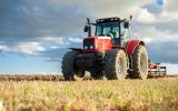 A person sitting in the cab of a red tractor in an open field. Though the sky is full of clouds, the sun shines.