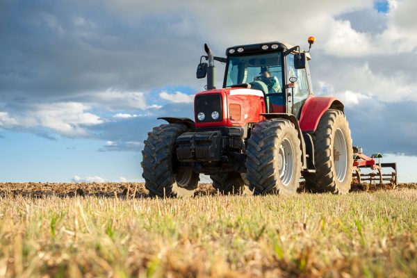 A person sitting in the cab of a red tractor in an open field. Though the sky is full of clouds, the sun shines.