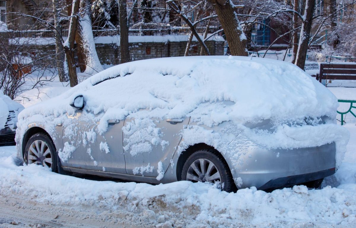 A silver car on the side of the road stuck in dirty snow and covered in a fresh sheet of snow.