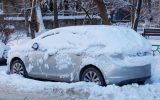 A silver car on the side of the road stuck in dirty snow and covered in a fresh sheet of snow.