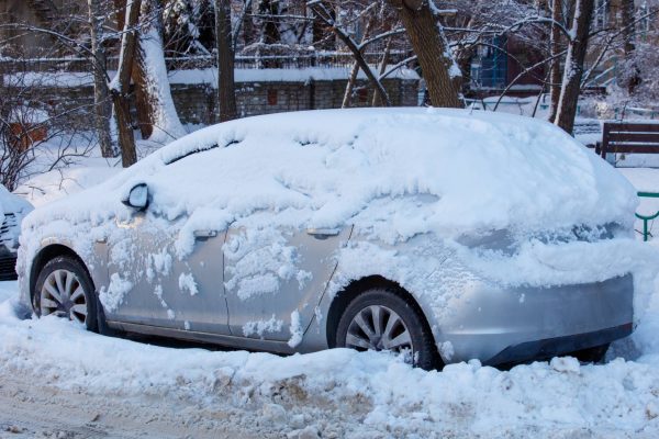 A silver car on the side of the road stuck in dirty snow and covered in a fresh sheet of snow.