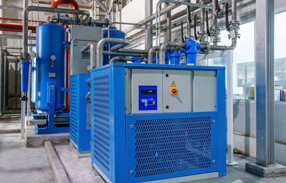 A couple of industrial air compressors with blue casing attached to metal piping on a concrete floor.