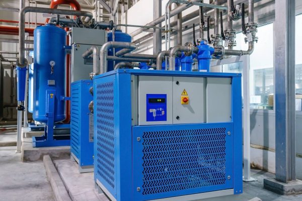 A couple of industrial air compressors with blue casing attached to metal piping on a concrete floor.