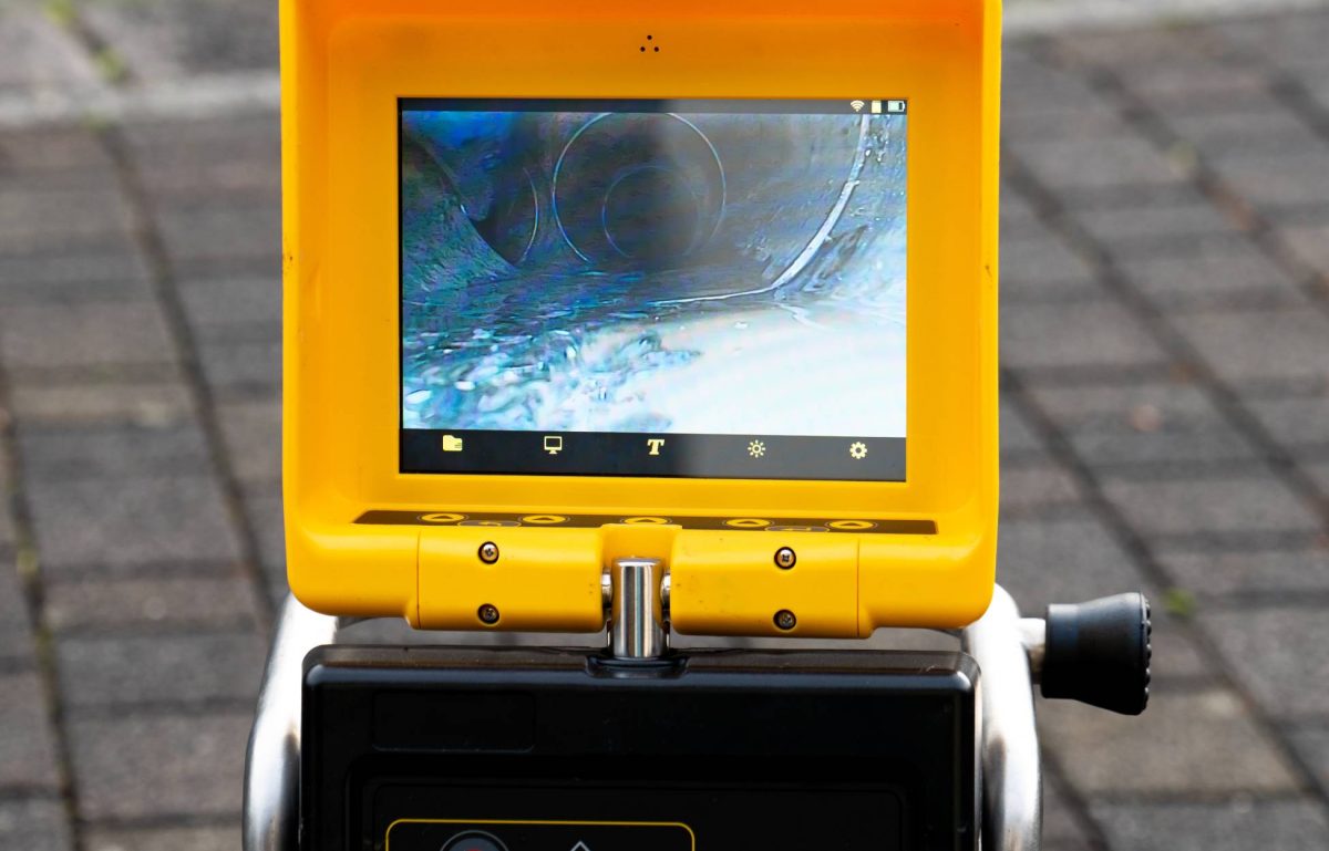 A close-up view shows a small screen on a piece of black and yellow equipment. The screen shows the inside of a drain.