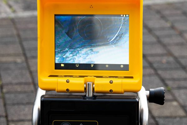 A close-up view shows a small screen on a piece of black and yellow equipment. The screen shows the inside of a drain.