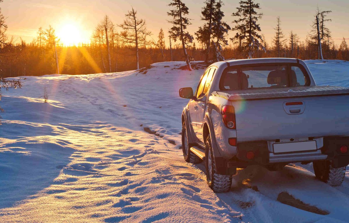 A white pick-up truck is parked in a snowy field with several pines as the sun rises above the horizon.