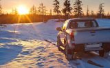 A white pick-up truck is parked in a snowy field with several pines as the sun rises above the horizon.