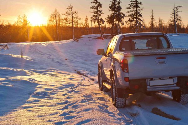 A white pick-up truck is parked in a snowy field with several pines as the sun rises above the horizon.