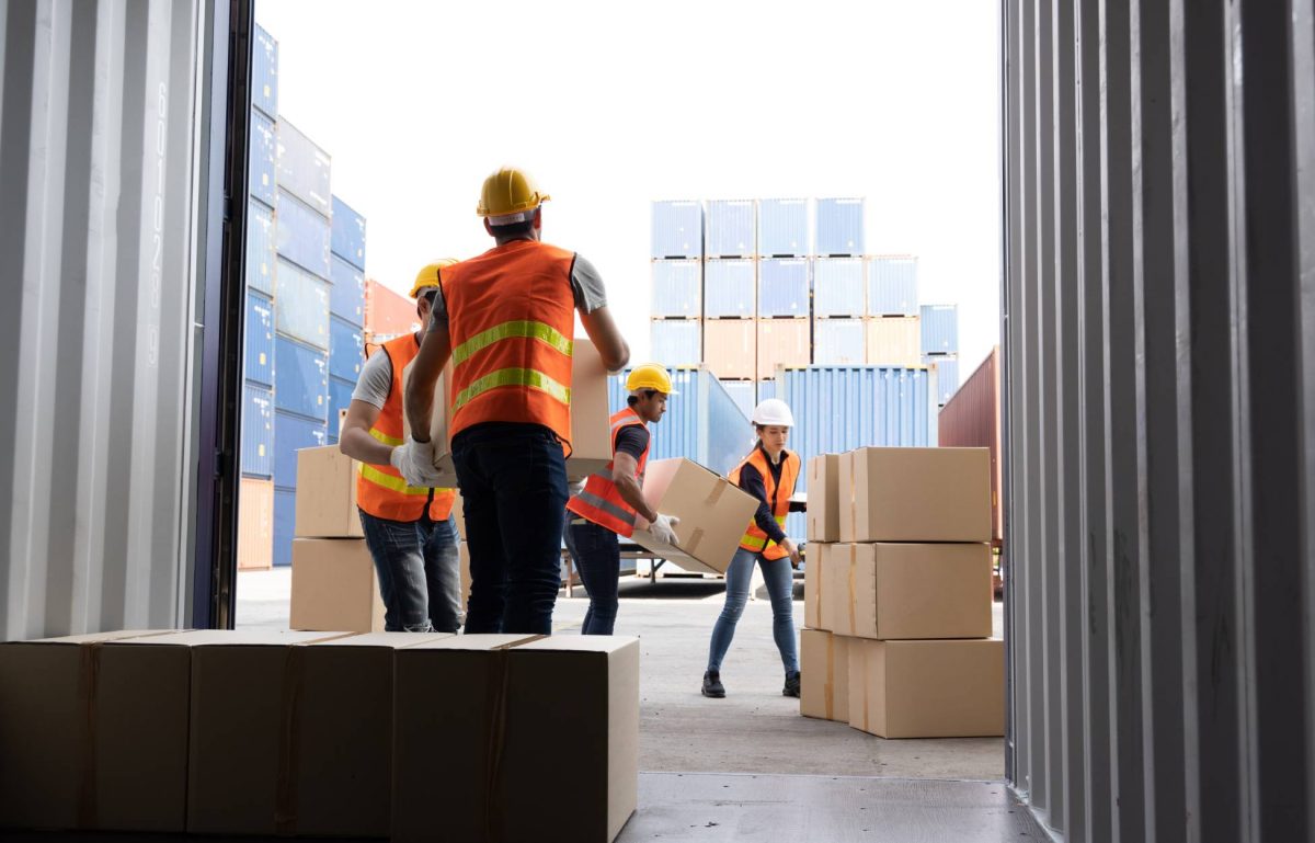 A crew of workers wearing orange and yellow vests and hard hats unload boxes from a shipping container.