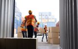 A crew of workers wearing orange and yellow vests and hard hats unload boxes from a shipping container.