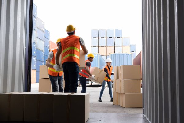 A crew of workers wearing orange and yellow vests and hard hats unload boxes from a shipping container.