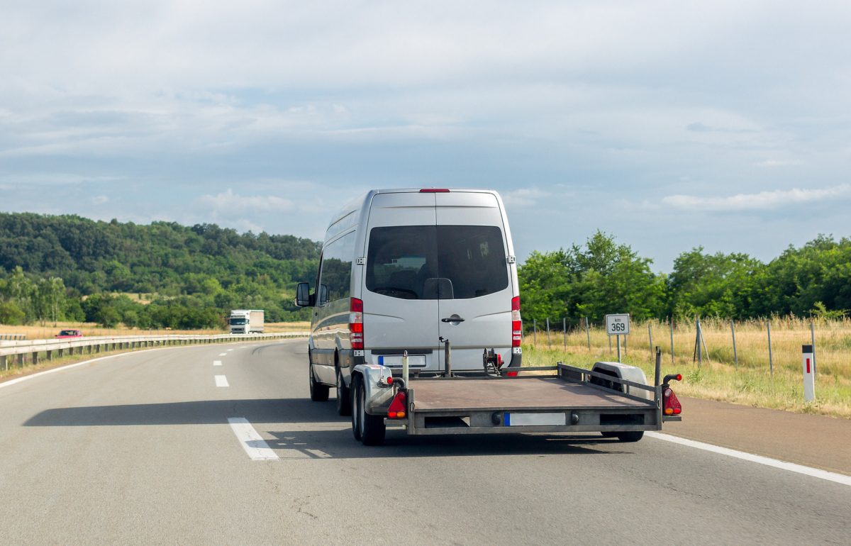 A white van drives down a highway while hauling an empty trailer. Two vehicles drive in the opposite direction.