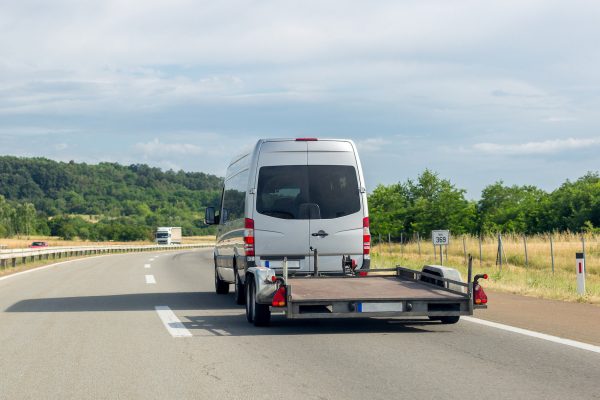 A white van drives down a highway while hauling an empty trailer. Two vehicles drive in the opposite direction.