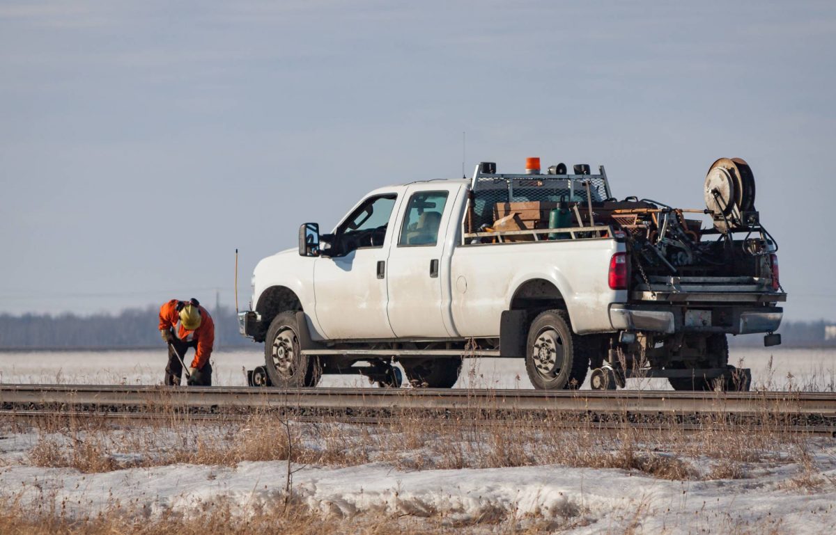 A work truck parked on the side of the road as a worker stands in front of it working on the road. The truck is white.