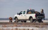 A work truck parked on the side of the road as a worker stands in front of it working on the road. The truck is white.