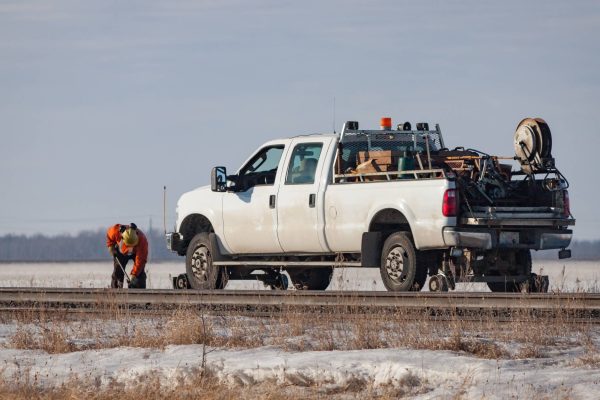 A work truck parked on the side of the road as a worker stands in front of it working on the road. The truck is white.