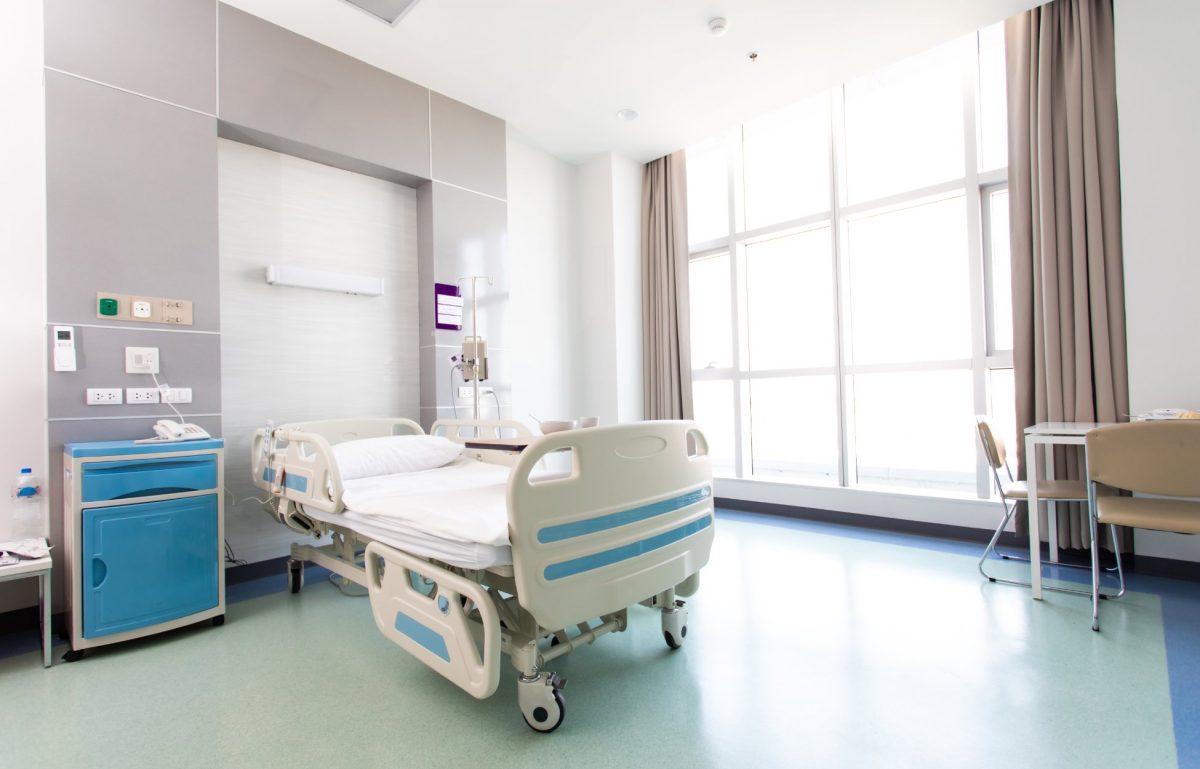 A hospital room contains an empty medical bed, a blue nightstand, a small table with tan chairs, and a large window.