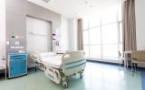 A hospital room contains an empty medical bed, a blue nightstand, a small table with tan chairs, and a large window.