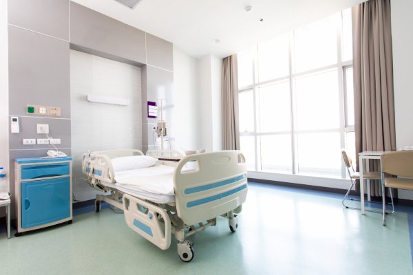 A hospital room contains an empty medical bed, a blue nightstand, a small table with tan chairs, and a large window.