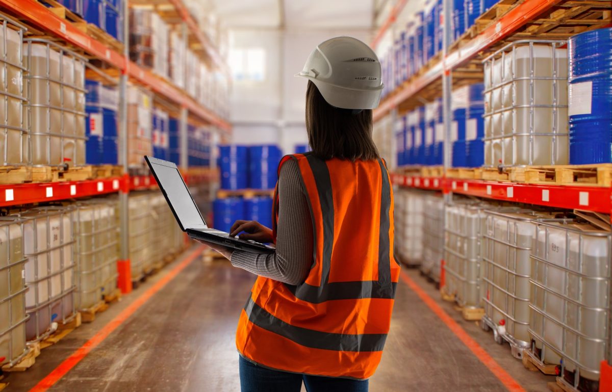 A woman wearing a white hard hat and an orange safety vest looks down a warehouse aisle holding a laptop.