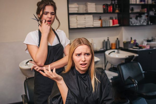 An upset salon client in a chair gestures angrily as her concerned stylist stands behind her with hair clippers.