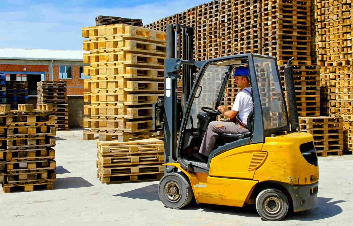 A person wearing a blue hard hat, white shirt, and gray pants drives a yellow forklift around piles of wooden pallets.