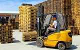 A person wearing a blue hard hat, white shirt, and gray pants drives a yellow forklift around piles of wooden pallets.