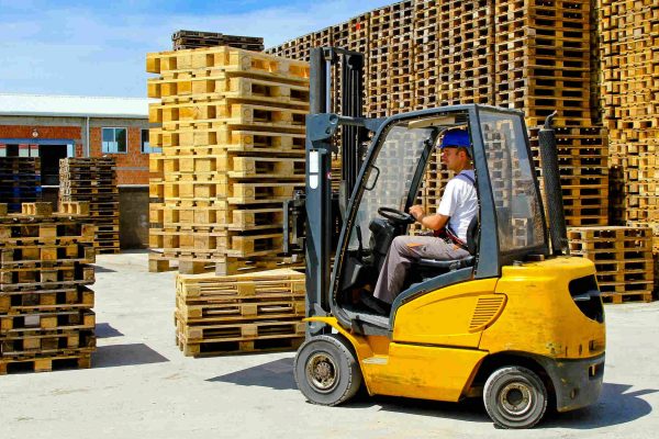 A person wearing a blue hard hat, white shirt, and gray pants drives a yellow forklift around piles of wooden pallets.