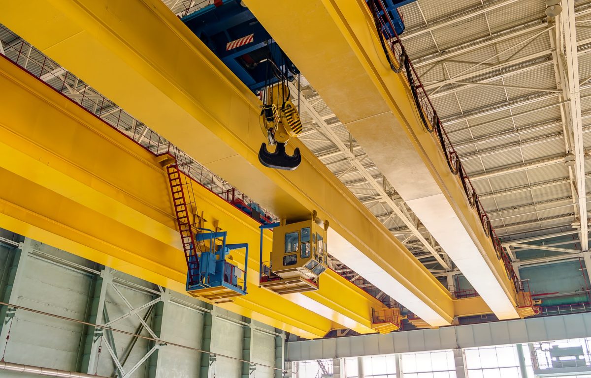 A ground-level point-of-view of a large, yellow overhead crane inside a factory featuring hooks, operator boxes, and more.