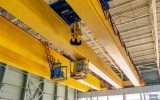 A ground-level point-of-view of a large, yellow overhead crane inside a factory featuring hooks, operator boxes, and more.