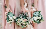 Three bridesmaids in light pink dresses stand together and hold large wedding bouquets of white flowers.