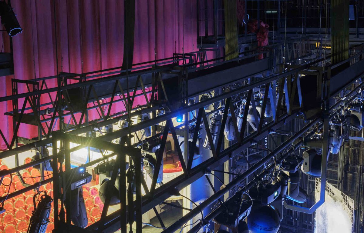 A grid of lights hangs above a lit stage. A red curtain and red theater seats are visible beyond the lighting equipment.