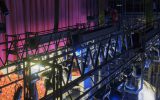 A grid of lights hangs above a lit stage. A red curtain and red theater seats are visible beyond the lighting equipment.