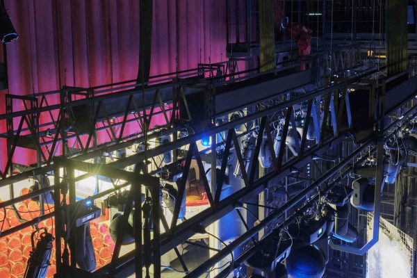 A grid of lights hangs above a lit stage. A red curtain and red theater seats are visible beyond the lighting equipment.