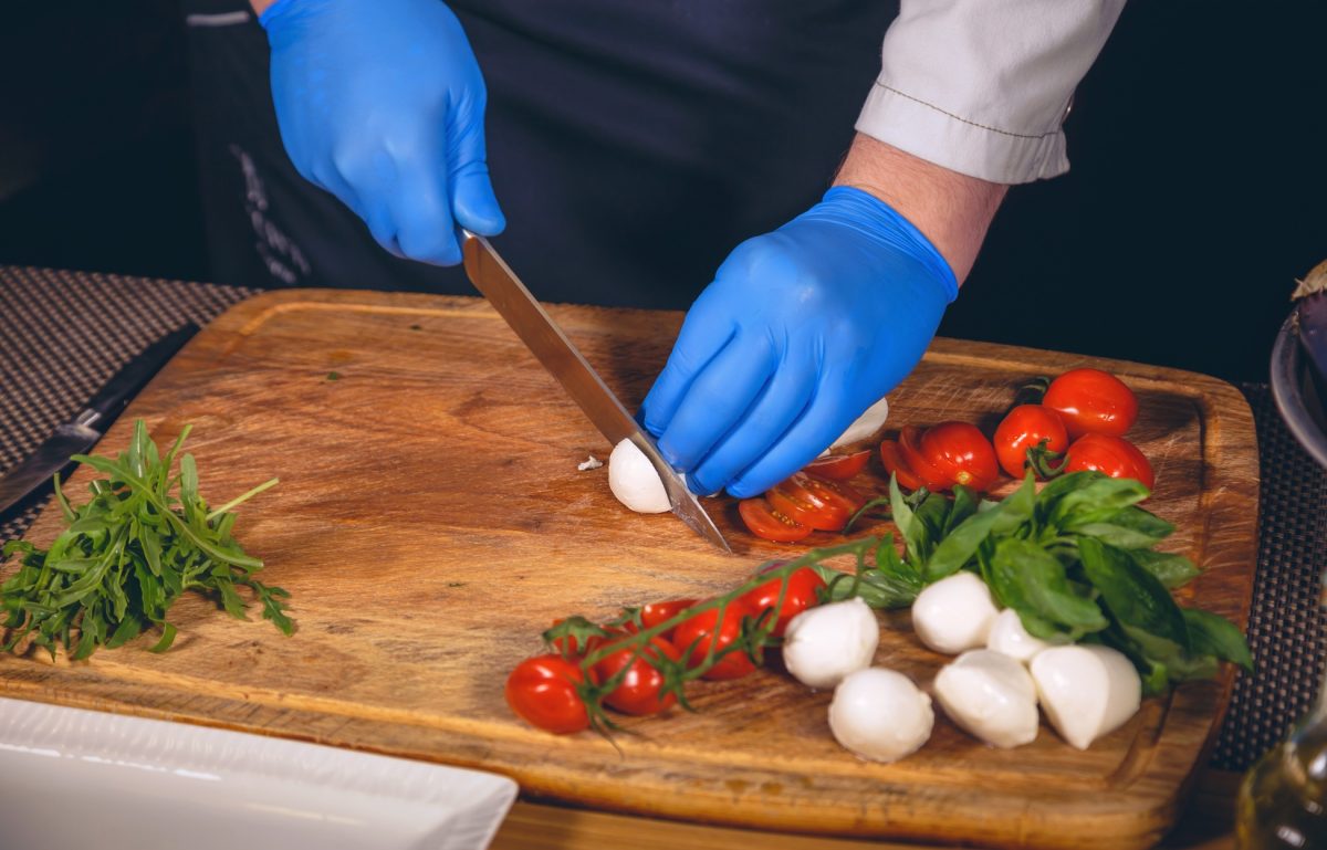 A close-up of a pair of hands in blue nitrile gloves using a chef's knife to prepare cheese, basil, and tomatoes.