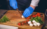 A close-up of a pair of hands in blue nitrile gloves using a chef's knife to prepare cheese, basil, and tomatoes.