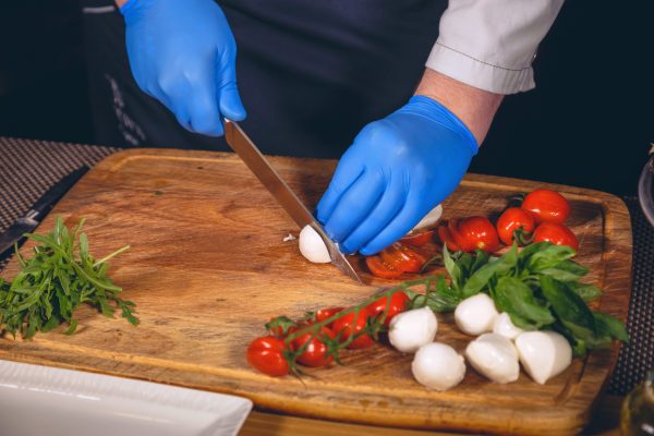 A close-up of a pair of hands in blue nitrile gloves using a chef's knife to prepare cheese, basil, and tomatoes.