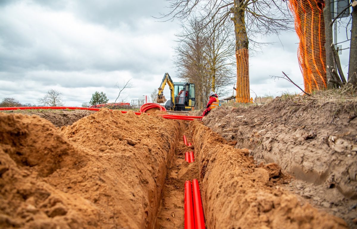 A trench containing partially buried bright red electrical lines. Two workers move additional lines above ground.