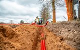 A trench containing partially buried bright red electrical lines. Two workers move additional lines above ground.