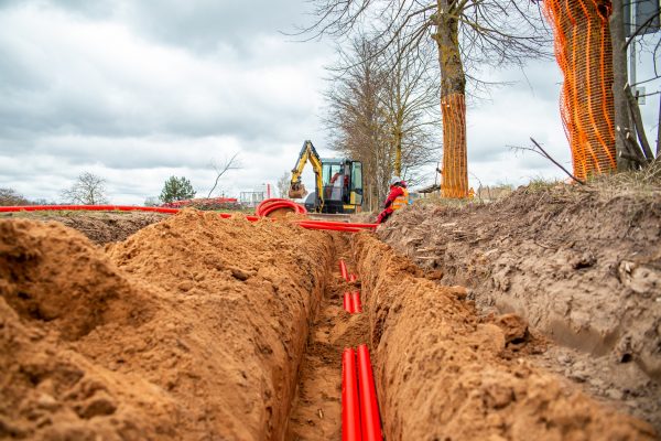 A trench containing partially buried bright red electrical lines. Two workers move additional lines above ground.