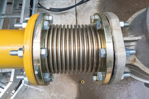 A top-down view of a metal expansion joint in an industrial setting. The expansion joint connects a yellow pipe to a silver one.